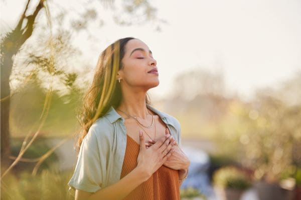 Donna in un prato con gli occhi chiusi e le mani sul cuore a simbolo di emozioni positive generate dalla respirazione modulata sul cuore